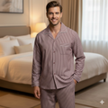 A male model smiling and standing in a cozy bedroom wearing a maroon and white striped wash-and-wear pajama set (CH-130). The long-sleeve shirt has a front pocket with perfectly aligned stripes, button closure, and white piping on the collar and cuffs.